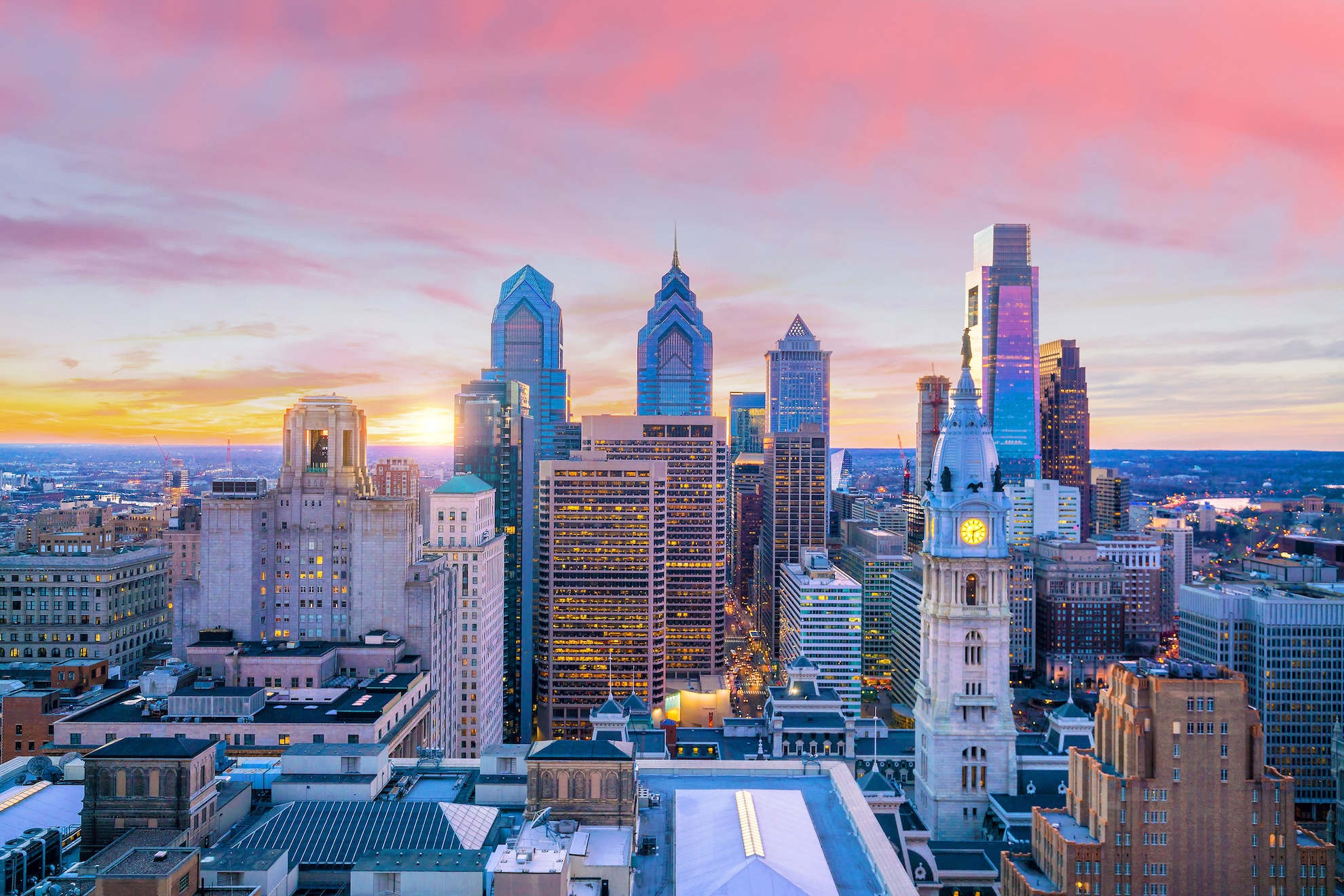 Downtown Philadelphia skyline at sunset with City Hall clock tower and skyscrapers under a pink and orange sky